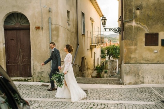 A bride and groom walk hand in hand down a cobblestone street in a quaint village, with the bride holding a bouquet of greenery and wearing a white dress, and the groom in a gray suit. by Justin Salem Meyer