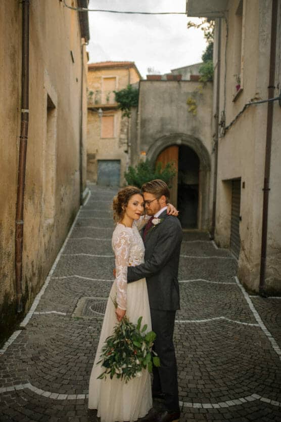 A bride and groom embrace on a narrow cobblestone street between old buildings. The bride wears a long-sleeve lace dress and holds a bouquet, while the groom wears a suit and looks at her lovingly. by Justin Salem Meyer