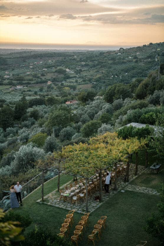 An outdoor dining area with a long table and chairs set under a vine-covered pergola overlooks a lush green valley at sunset. A few people stand nearby, and hills stretch into the distance under a colorful sky. by Justin Salem Meyer