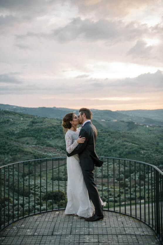 A bride and groom embrace on a circular metal balcony overlooking rolling green hills at sunset, with dramatic clouds in the sky. by Justin Salem Meyer