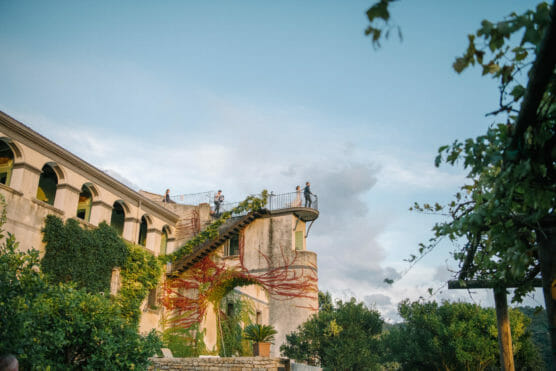 A rustic villa covered in greenery and red vines, with people standing on a rooftop terrace, overlooks lush gardens under a blue sky with scattered clouds. by Justin Salem Meyer