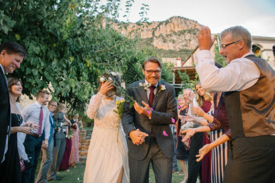A newlywed couple walks outside as guests throw flower petals at them, smiling and celebrating. The bride covers her face while the groom laughs. Mountains and greenery are visible in the background. by Justin Salem Meyer