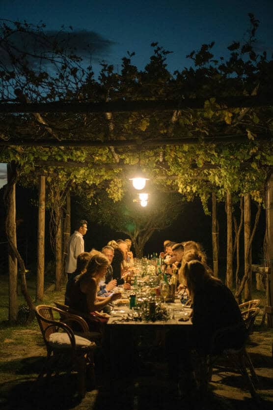 People are gathered around a long outdoor dinner table under string lights and vine-covered pergola at night, sharing a meal and conversation in a relaxed, festive atmosphere. by Justin Salem Meyer