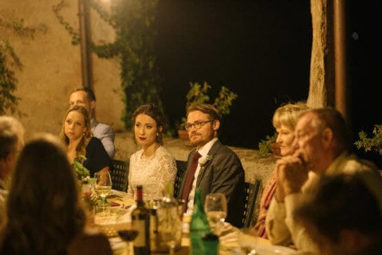 A bride and groom sit together at a warmly lit dinner table outdoors, surrounded by guests. The atmosphere is intimate and festive, with wine glasses and plates on the table. Everyone appears to be attentively listening. by Justin Salem Meyer