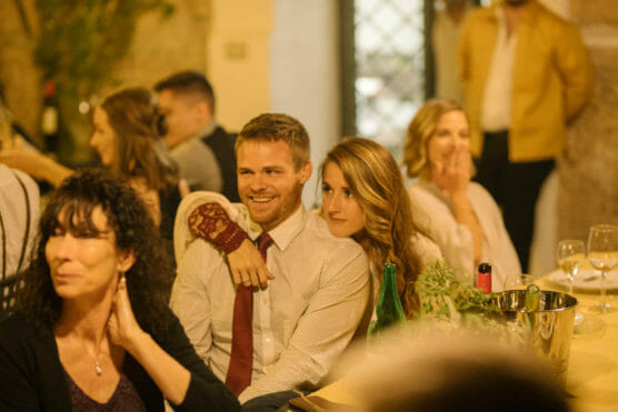 A smiling couple sits closely together at a warmly lit dinner event, surrounded by other guests. The woman has her arm around the man, and wine glasses and bottles are on the table in front of them. by Justin Salem Meyer