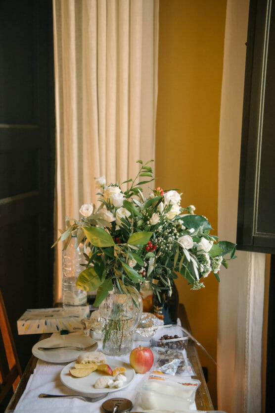 A glass vase with white flowers and greenery sits on a wooden table by a window, surrounded by a plate of food, an apple, bottled water, and a few food containers. Light filters through beige curtains. by Justin Salem Meyer