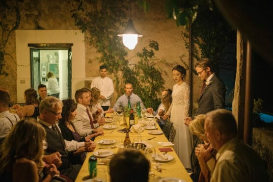 A bride and groom stand at the head of a long dinner table, surrounded by seated guests in a warmly lit, rustic room with ivy-covered walls. Chefs are visible in the background through an open doorway. by Justin Salem Meyer