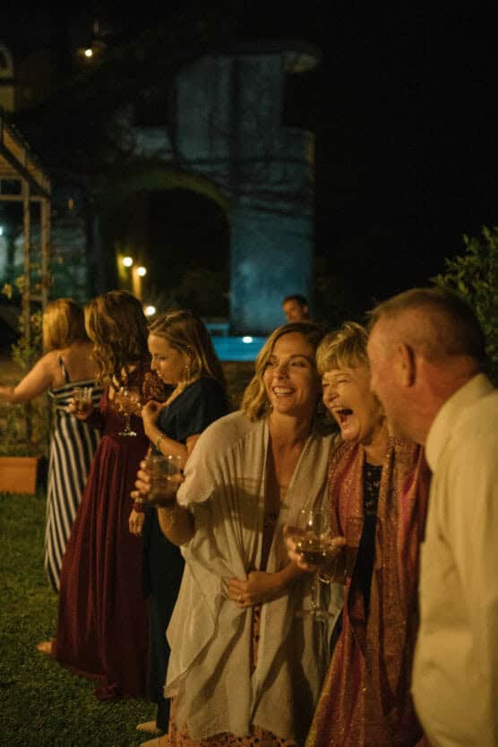 A group of people dressed formally stand outdoors at night, smiling and laughing while holding drinks. The atmosphere is festive, with warm lighting and greenery in the background. by Justin Salem Meyer