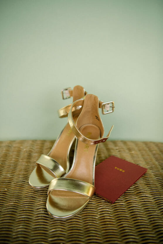 A pair of gold high-heeled sandals with ankle straps is placed on a woven surface next to a small red booklet with gold lettering. by Justin Salem Meyer
