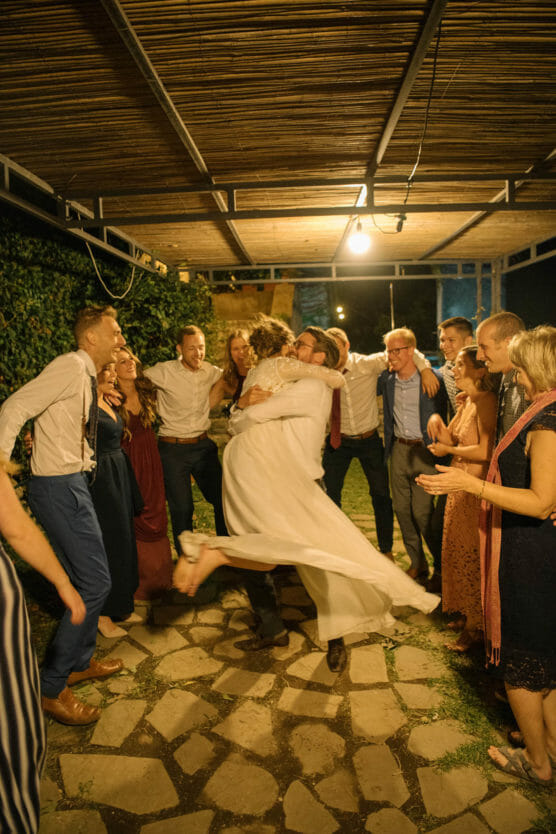 A joyful group of people celebrate outdoors at night; one person in a white dress is lifted up in a spinning hug by another, while friends around them laugh, clap, and smile under string lights. by Justin Salem Meyer