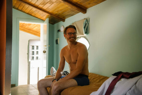 A smiling man wearing glasses and shorts sits shirtless on a bed in a sunlit room with wooden ceiling, an open door behind him, and clothes and an iron visible nearby. by Justin Salem Meyer