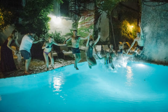 A group of people holding hands jump into a lit swimming pool at night, splashing water. Some are mid-air, while others are already in the pool. Trees and illuminated walls surround the pool area. by Justin Salem Meyer