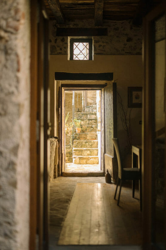 A cozy, rustic hallway with stone walls and wooden beams leads to an open doorway, revealing stone steps outside illuminated by warm sunlight. A chair and table are visible on the right side of the hallway. by Justin Salem Meyer