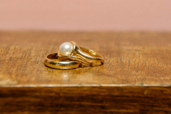 Two gold rings resting on a wooden surface; one is a plain band, and the other features a single white pearl set in a simple gold setting. by Justin Salem Meyer