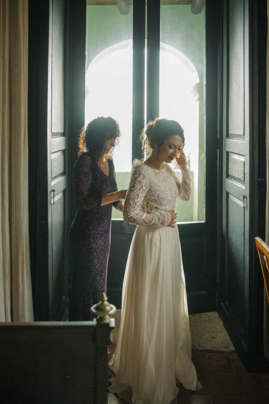 A woman in a long-sleeved white wedding dress stands by a sunlit doorway while another woman in a dark lace dress helps her adjust the gown. Soft light filters through the open doors behind them. by Justin Salem Meyer