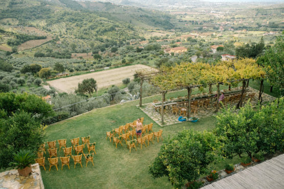 Rows of empty wicker chairs are arranged on a grassy hilltop for an outdoor event, overlooking a scenic valley with green fields, distant houses, and rolling hills. A pergola covered in vines provides shade nearby. by Justin Salem Meyer