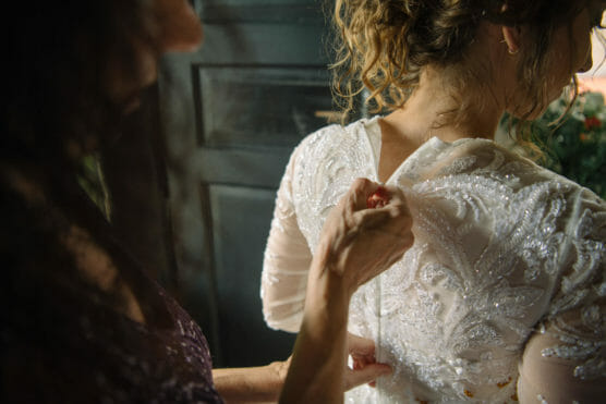 A close-up of a woman buttoning the back of a brides lace wedding dress, focusing on the intricate details of the gown and the hands assisting, with soft natural light illuminating the scene. by Justin Salem Meyer