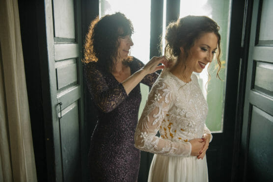 A woman in a lace wedding dress smiles as another woman helps fasten her dress, standing together by a window with soft natural light shining in. by Justin Salem Meyer