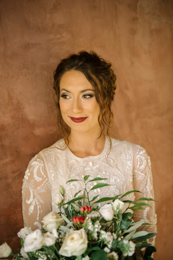 A woman in a white lace dress with wavy brown hair holds a bouquet of white and red flowers, standing against a textured brown background and looking to the side with a soft smile. by Justin Salem Meyer
