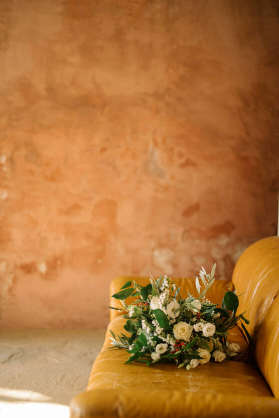 A bouquet of white flowers with green leaves rests on a yellow couch against a textured, warm-toned, peach-colored wall. by Justin Salem Meyer