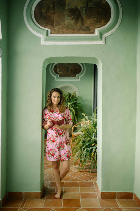 A woman in a pink floral robe stands barefoot in a green hallway with terracotta floor tiles, holding a book and looking at the camera. There are lush green plants and decorative wall art in the background. by Justin Salem Meyer