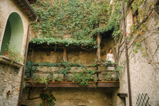 A woman in a white dress stands on a rustic balcony covered in green vines, surrounded by old stone walls and lush plants in a charming courtyard. by Justin Salem Meyer