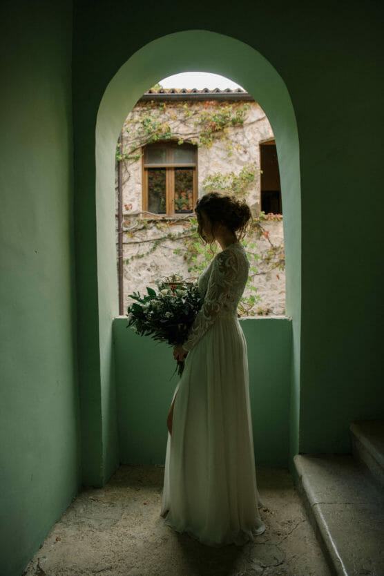 A woman in a long-sleeved white dress stands in an arched doorway, holding a bouquet of greenery, with soft light illuminating her silhouette and an old stone building visible outside. by Justin Salem Meyer