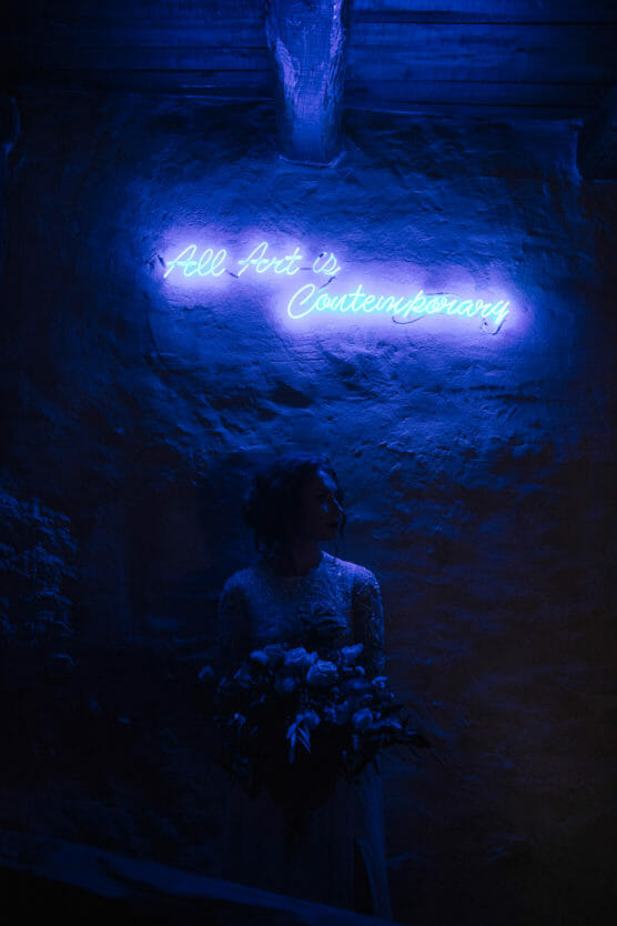 A person in a white dress holding a bouquet stands under a glowing blue neon sign on a textured wall that reads, All Art is Contemporary. by Justin Salem Meyer