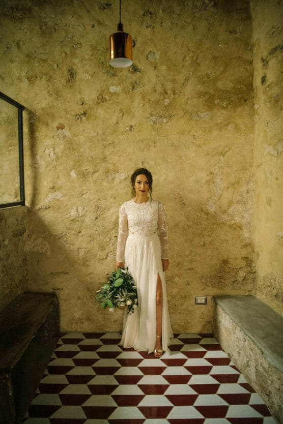 A bride in a long-sleeved white gown holds a bouquet of greenery, standing against a textured yellow wall with a tiled red-and-white floor and a hanging light above. by Justin Salem Meyer