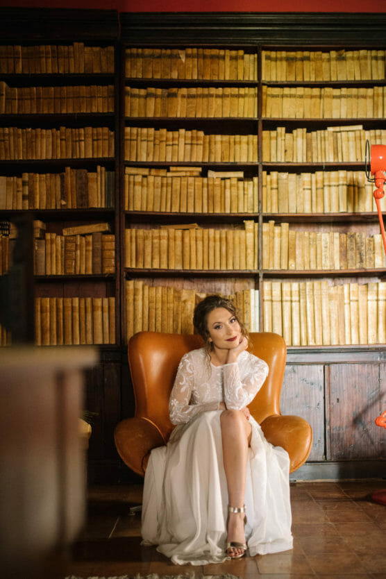 A woman in a white dress sits in a brown armchair, resting her chin on her hand, in front of a large bookshelf filled with old books. The setting is warmly lit and elegant. by Justin Salem Meyer