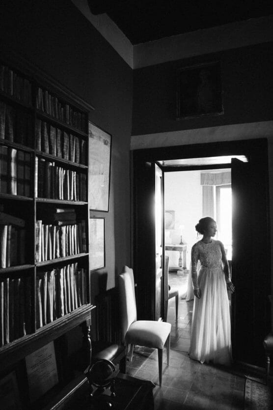 A woman in a long, elegant dress stands in a doorway, looking to the side. The room contains tall bookshelves filled with books and is lit softly, creating a classic, vintage atmosphere. The image is in black and white. by Justin Salem Meyer