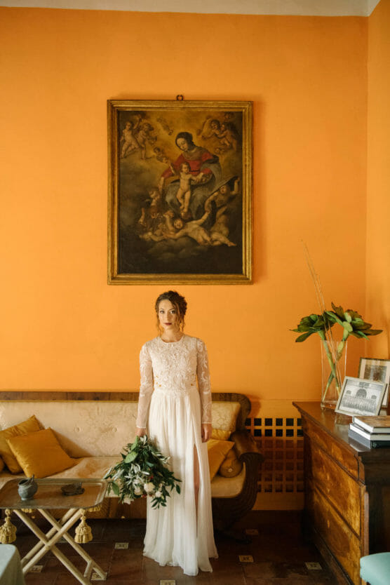 A woman in a long, white lace dress holding a bouquet stands in a warm, orange-toned room with a painting above a beige couch, a wooden table, and a sideboard with plants and framed photos. by Justin Salem Meyer