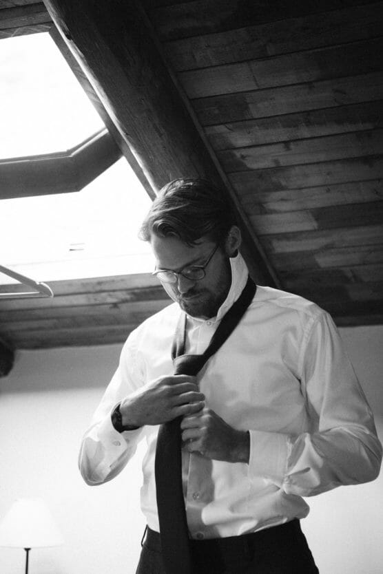 A man wearing glasses and a white shirt stands indoors, looking down as he adjusts a necktie. The room has a wooden ceiling with exposed beams and a skylight above him. The atmosphere appears calm and focused. by Justin Salem Meyer
