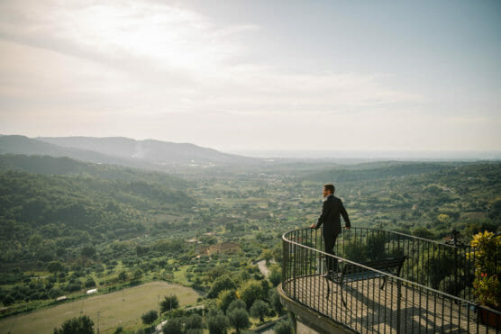 A man in a suit stands on a balcony overlooking a vast, scenic valley filled with green hills and trees under a bright sky. by Justin Salem Meyer