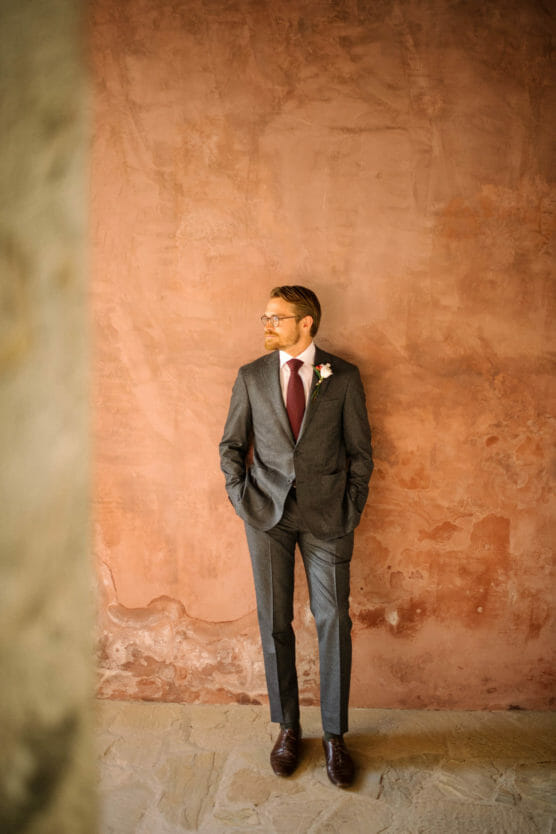 A man in a gray suit with a red tie and boutonniere stands with hands in pockets against a textured, reddish-brown wall, gazing to the side. by Justin Salem Meyer