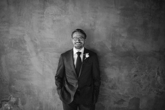 A man in a suit with a boutonnière smiles and stands against a textured wall with his hands in his pockets. The photo is in black and white. by Justin Salem Meyer