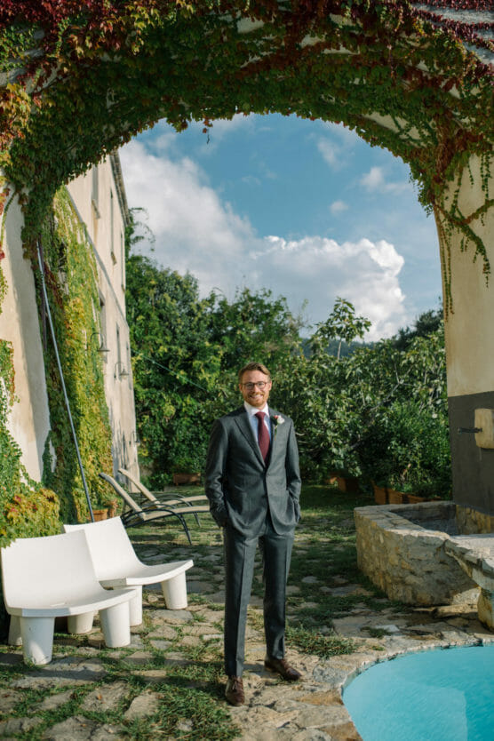 A man in a gray suit stands by a pool under a vine-covered stone archway, with a building and lush greenery in the background. Two white chairs are beside him, and the sky is partly cloudy. by Justin Salem Meyer