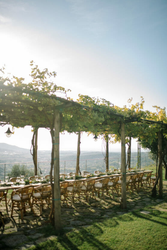 A long outdoor dining table with wooden chairs sits under a pergola covered in green vines, overlooking a scenic landscape with hills and a clear blue sky. Sunlight filters through the leaves, casting dappled shadows. by Justin Salem Meyer