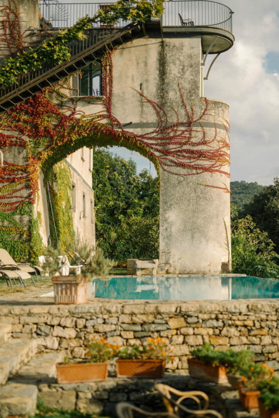 A rustic stone building with an arched wall covered in red and green vines stands beside a clear swimming pool, surrounded by lush greenery and outdoor lounge chairs under a partly cloudy sky. by Justin Salem Meyer