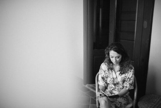 A woman with wavy hair, wearing a floral robe, sits on a chair near a door, looking down thoughtfully, in a softly lit room. The left side of the image is bright and out of focus. by Justin Salem Meyer