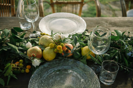 A rustic wooden table set with clear glass plates, wine glasses, and water glasses, decorated with a lush garland of greenery, lemons, pomegranates, and white flowers. Wicker chairs are visible in the background. by Justin Salem Meyer