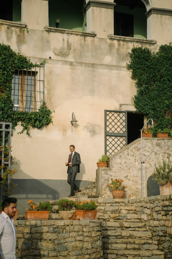 A man in a suit stands on stone steps by a rustic building covered with ivy and potted plants. Another person in a white shirt is partially visible at the bottom left. Sunlight casts warm tones on the scene. by Justin Salem Meyer