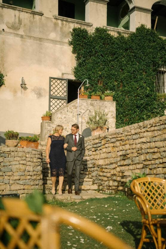 A woman in a navy lace dress and a man in a suit with a red tie walk arm-in-arm down stone steps beside a vine-covered wall, with sunlight illuminating the outdoor setting. by Justin Salem Meyer