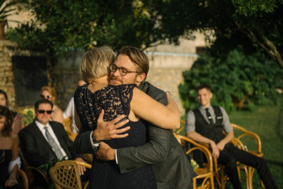 A man in a suit hugs a woman in a navy lace dress at an outdoor event, with other guests seated on wooden chairs and greenery in the background. by Justin Salem Meyer