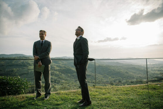Two men in suits stand on a grassy hilltop overlooking a scenic valley with mountains in the distance, under a partly cloudy sky near sunset. One man is holding a woven folder. by Justin Salem Meyer