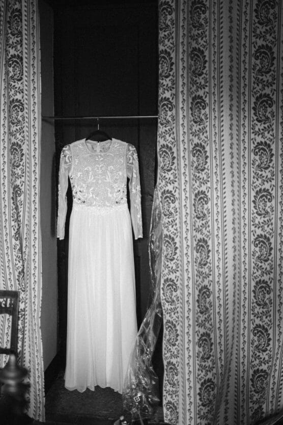 A long-sleeved lace wedding dress hangs in an open closet between patterned curtains. The elegant gown features intricate embroidery on the bodice and a flowing, sheer skirt. The image is in black and white. by Justin Salem Meyer