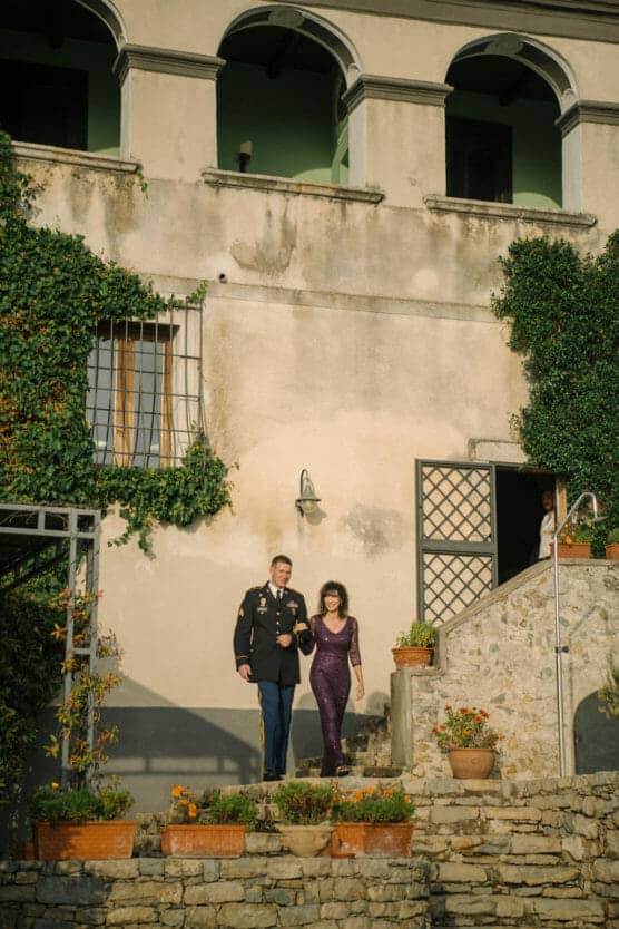 A man in a formal military uniform and a woman in a sparkly purple dress walk arm-in-arm down stone steps outside a sunlit, ivy-covered building with potted plants and arched balconies. by Justin Salem Meyer