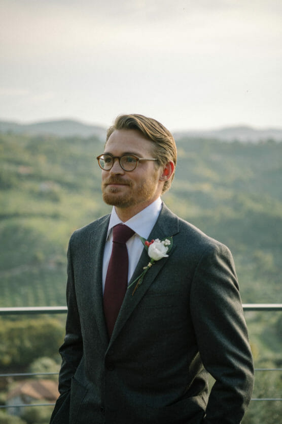 A man wearing glasses, a dark suit, maroon tie, and a white rose boutonniere stands outdoors with green hills and a soft, hazy sky in the background. by Justin Salem Meyer