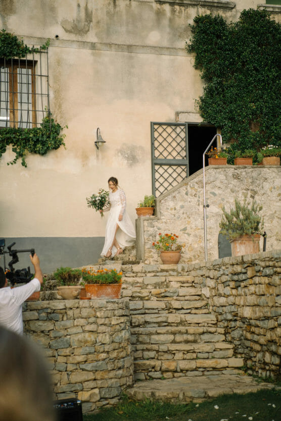 A bride in a white dress walks down stone steps outside a rustic building with ivy and potted plants, holding a bouquet of greenery. A person in the foreground is filming the scene. by Justin Salem Meyer