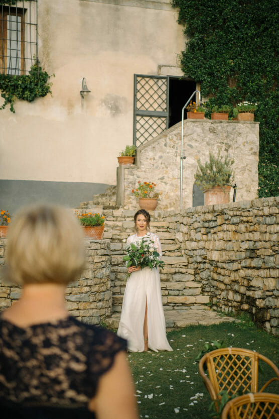 A bride in a white dress holding a bouquet walks down stone steps outdoors, while a seated guest with short blonde hair watches her. The scene is set in a rustic courtyard with plants and stone walls. by Justin Salem Meyer